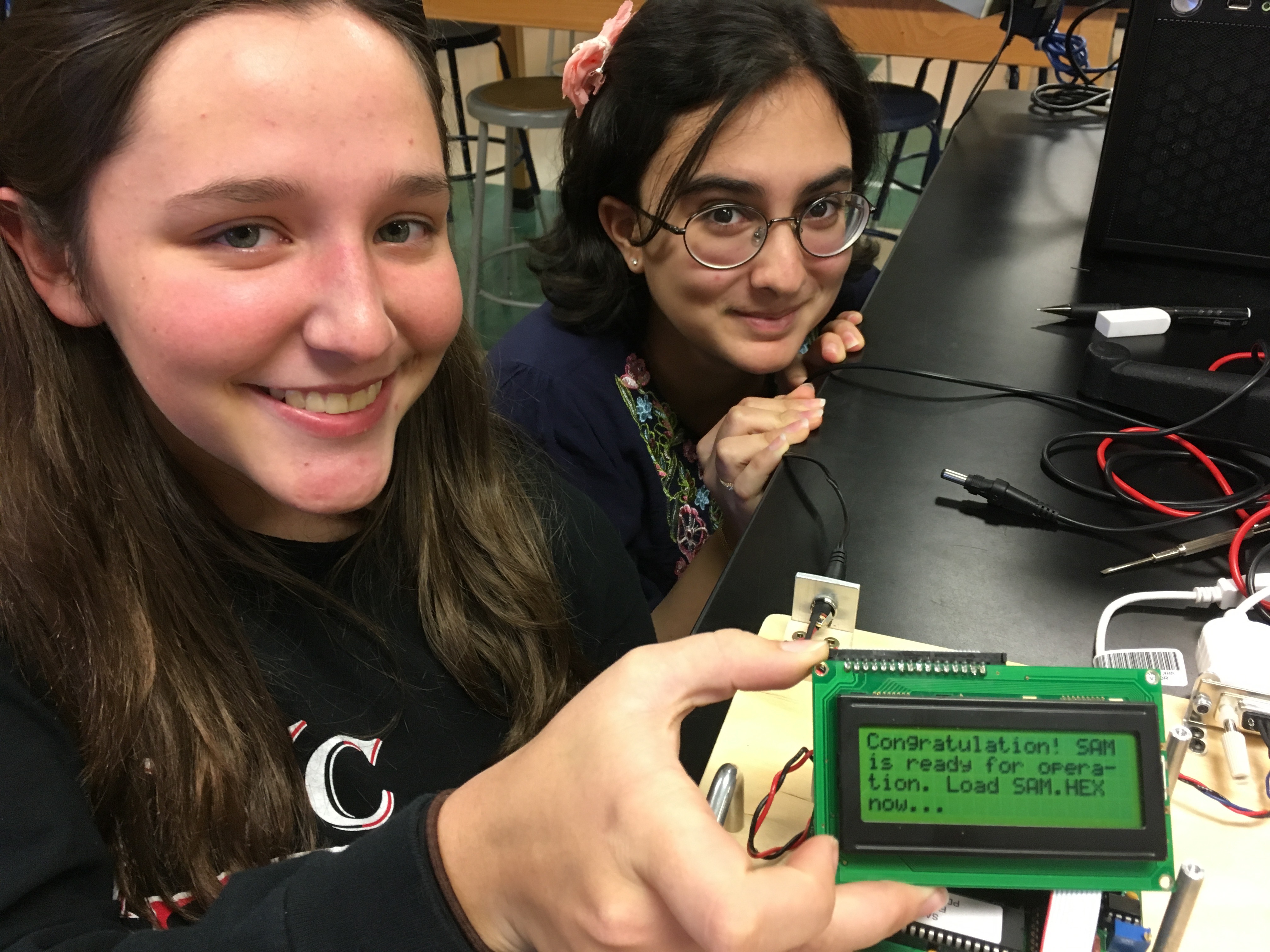 Two young female students sit with a small space instrument they built.