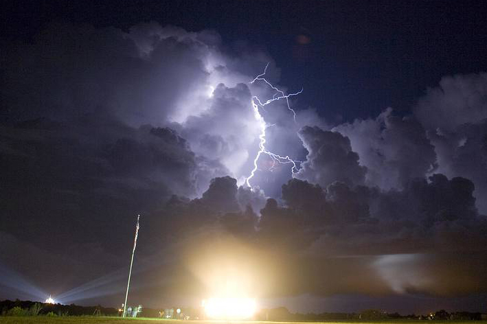 White-blue lightning appears out of a black cloud and hits the ground with a bright yellow burst.