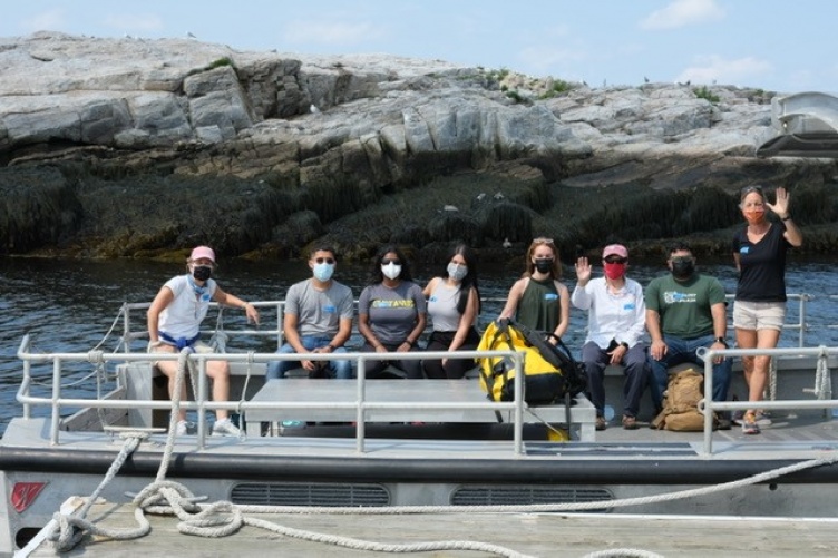 Students aboard a boat next to a rocky island wave at the camera.