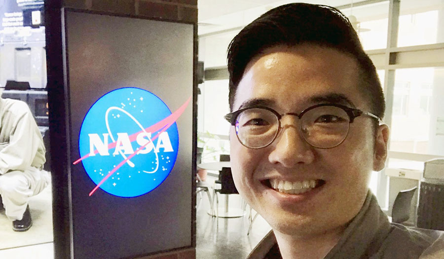 Eunsang Cho next to the NASA logo sign inside his office building. Eunsang standing next to NASA logo sign inside building.
