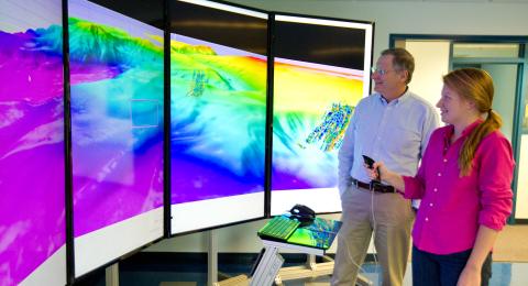 Man and woman stand with hand-held controller near large, four-paneled computer screen of colorful underwater maps.