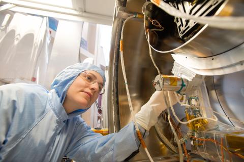 Woman wearing glasses and a blue Tyvek suit and white latex gloves inspects silver space instrument with wires coming out of it.