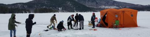 People in winter clothes stand on ice-covered pond lowing acoustic equipment under the water. 