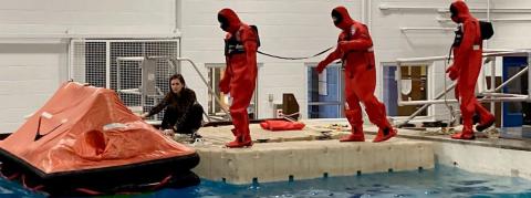 Three people in red survival suits approaching raft in tank. 