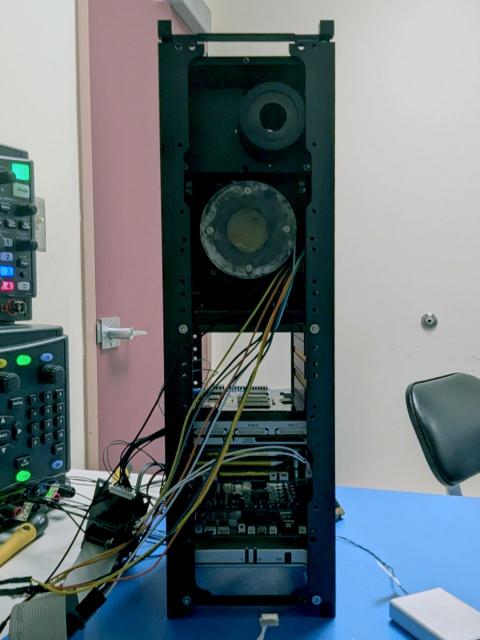 Photo of a tall satellite cube sitting on a desk with wiring. 