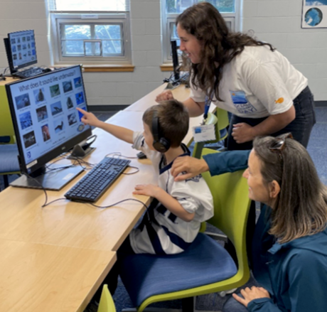 Photo of boy pointing at monitor with two women behind him. 