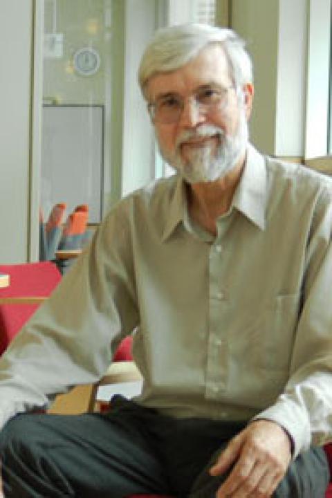 An older man with white hair, beard, mustaches and glasses sits on a chair and gives a small smile for the camera.
